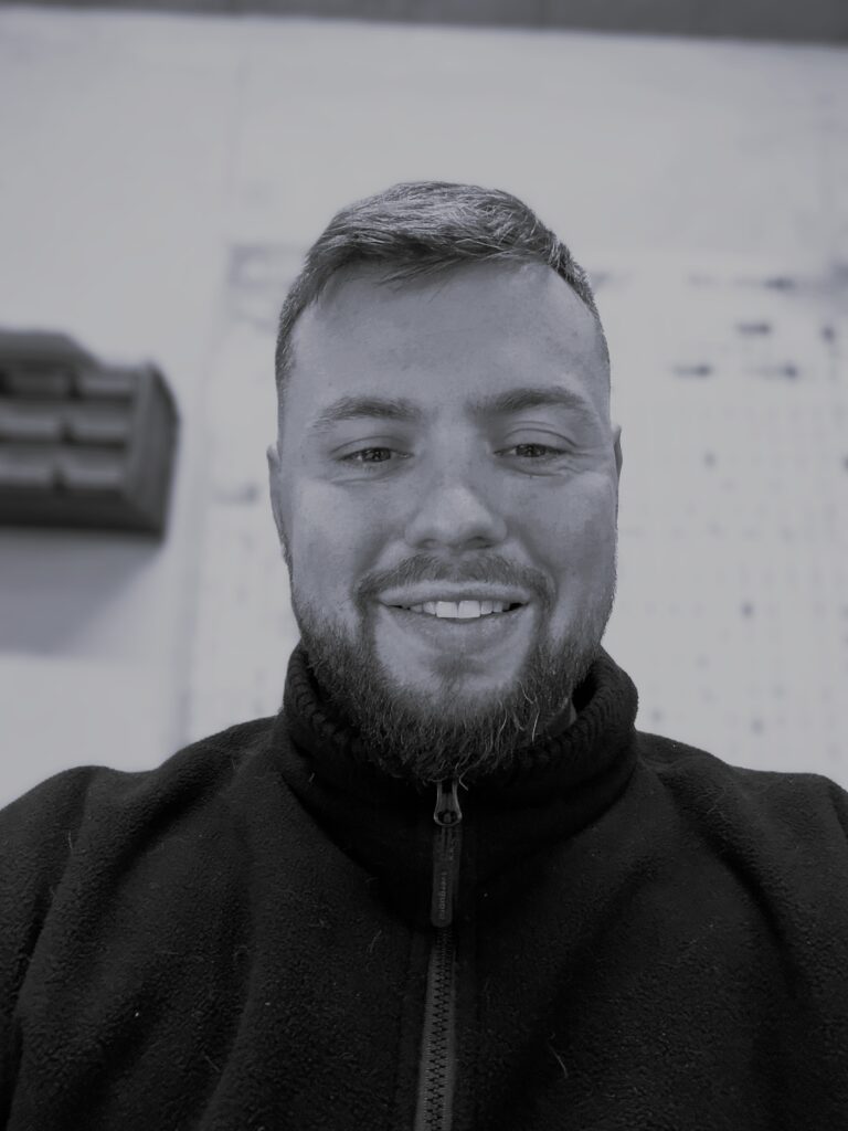 Homme Barbu Souriant en Polaire Noire Portrait en noir et blanc d'un homme souriant avec une barbe, portant une veste polaire noire zippée. Le zipper indique 'overguard'.
