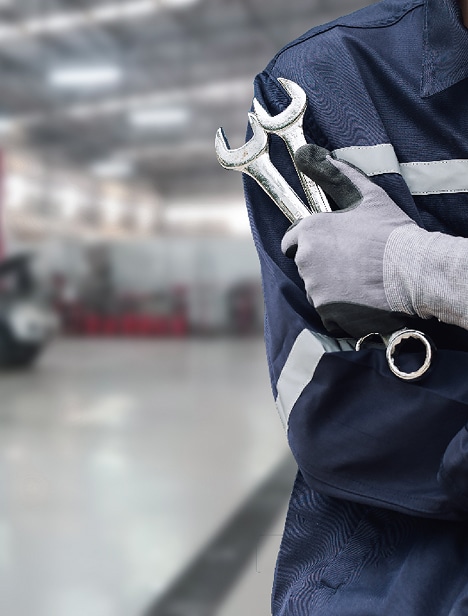 Mécanicien avec une combinaison bleue à bandes réfléchissantes tenant deux clés plates dans un atelier de réparation automobile flou.